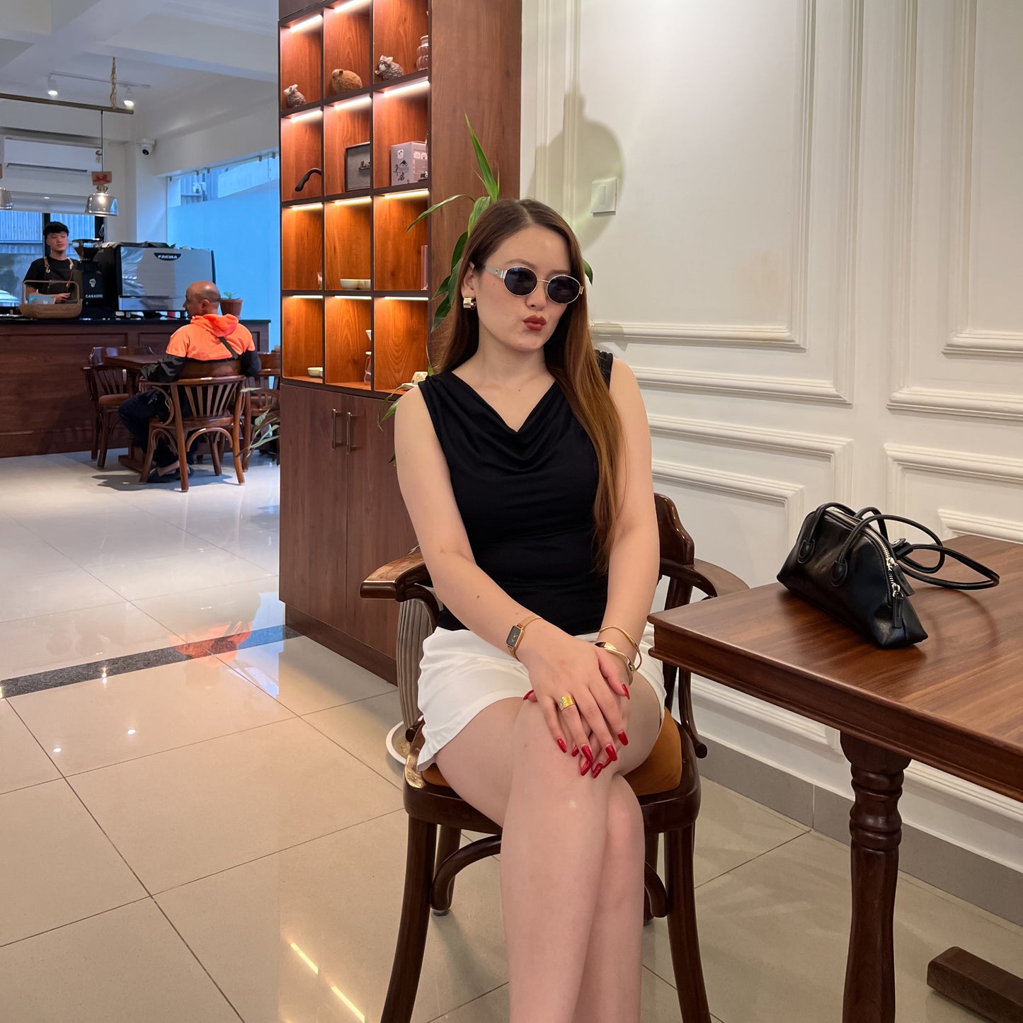 A woman wearing a black sleeveless cowl-neck top and a white skirt sits in a cafe setting, posing in front of a wooden display cabinet and table.