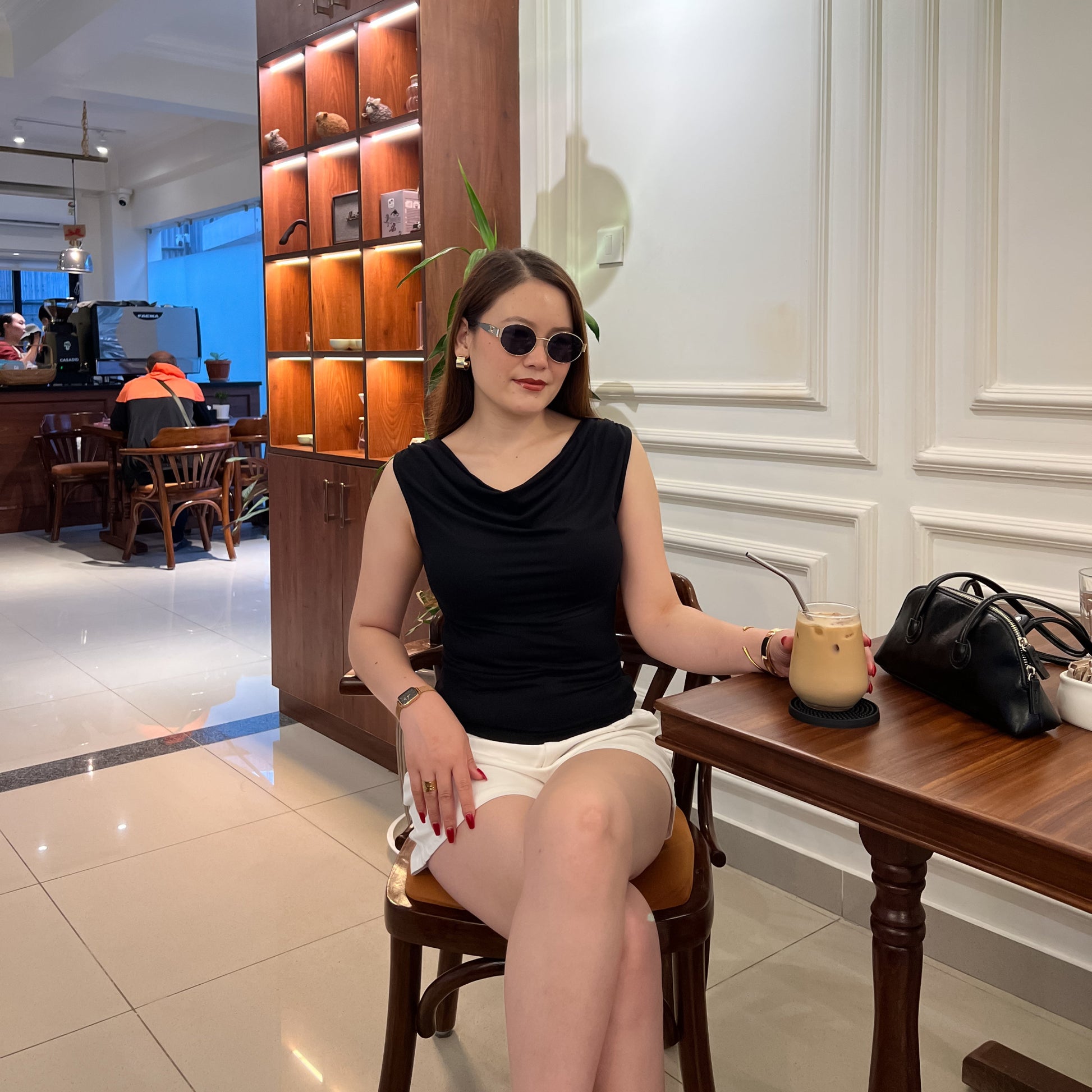 A woman wearing a black sleeveless top and white shorts sits at a wooden table in a cafe, holding a drink. She is wearing round sunglasses, and a black handbag rests on the table next to her.
