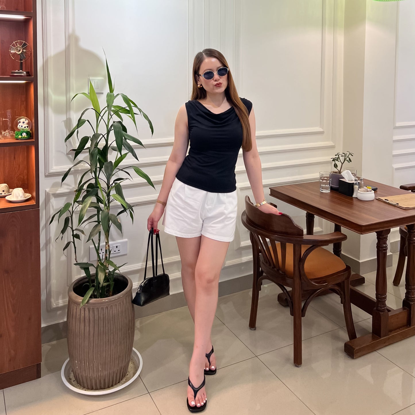 A woman wearing a black Grace Cowl Neck Sleeveless top in medium size by Laconic Fashion Nepal, white shorts, black sandals, and sunglasses stands in a cafe-like interior with a wooden table, chairs, potted plant, and hanging green lamps.