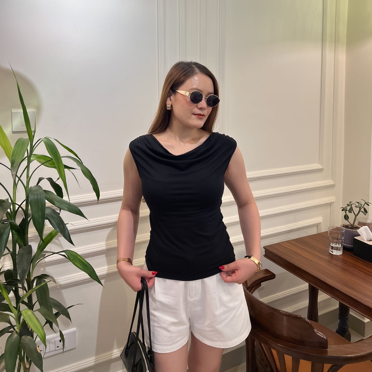 A woman wearing a black cowl-neck sleeveless top by Laconic Fashion Nepal and white shorts stands indoors in a room with white paneled walls, a green pendant light, a plant, and a wooden table.