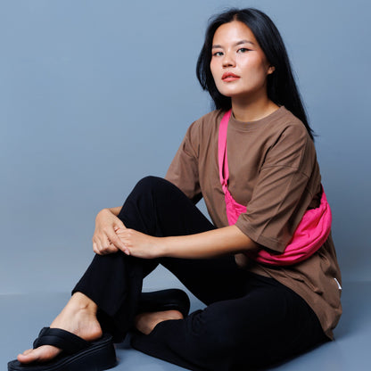 A model sits cross-legged on a blue-gray studio floor wearing Laconic Fashion Nepal brown oversized T-shirt, black trousers, black platform sandals, and a bright pink sling bag across her torso. Laconic Fashion Nepal Straight Leg Trousers ideal for Summer